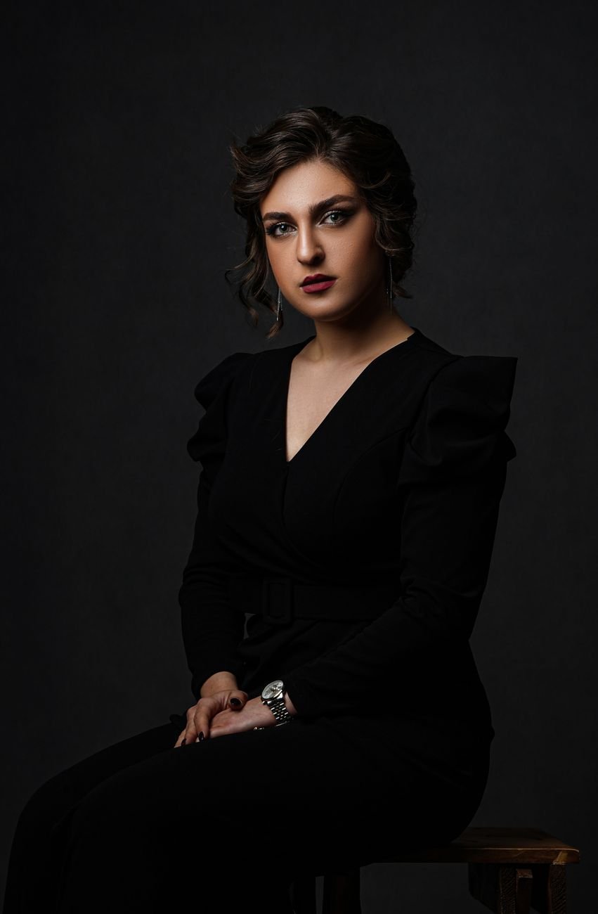 Portrait of a stylish woman in a black dress, sitting in a studio with a dark background.